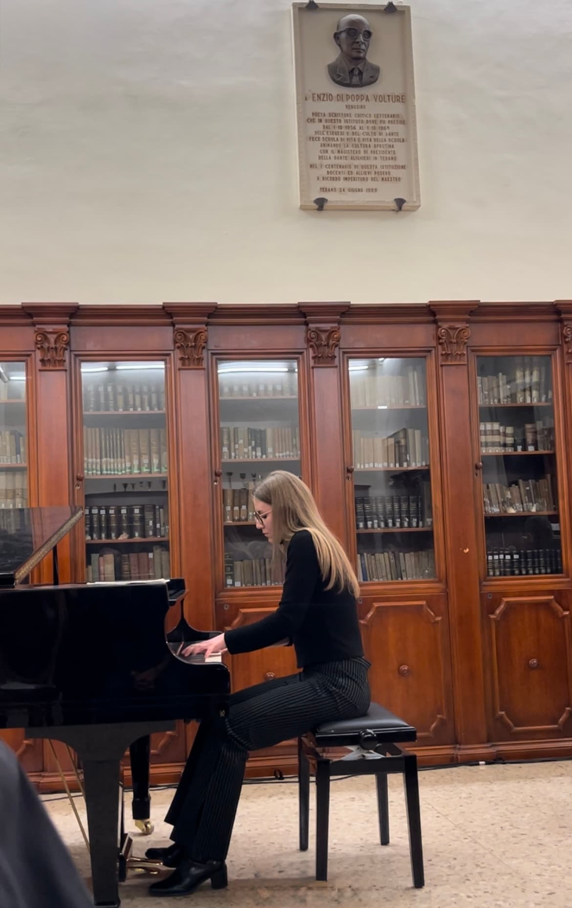 Una ragazza bionda suona un pianoforte a coda nero davanti a grandi librerie in legno.